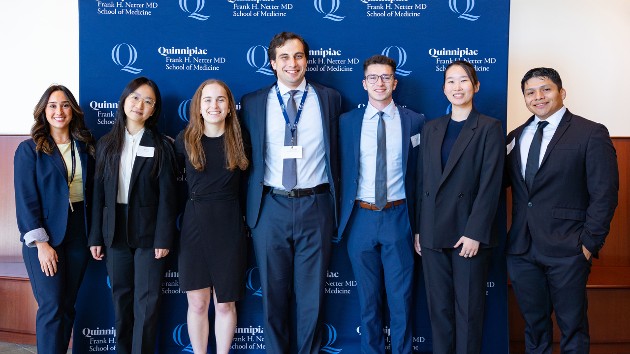 Seven students pose for a photo in front of Quinnipiac backdrop.