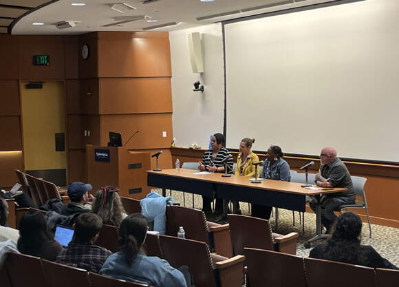Moderators and panelists speak at the panel in front of a crowd of people.
