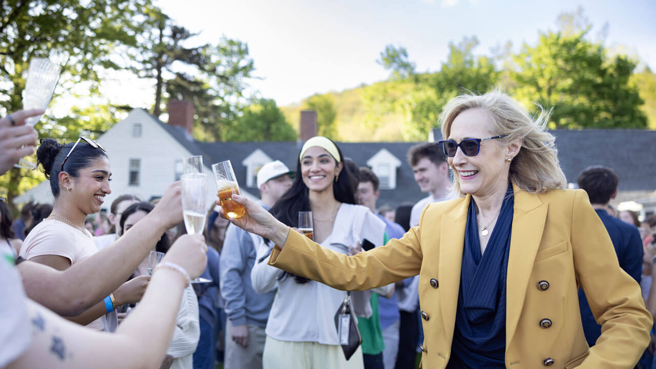 President Marie Hardin, wearing a yellow suit and sunglasses, smiles and raises a drink in a toast with students gathered outdoors at Quinnipiac University’s 2026 Class Toast