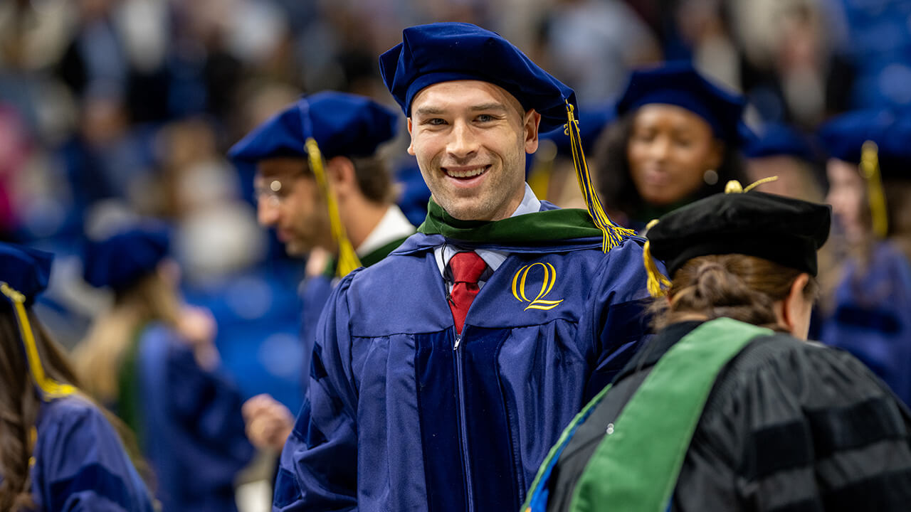 Graduate smiles during ceremony around peers, as they stand in the background