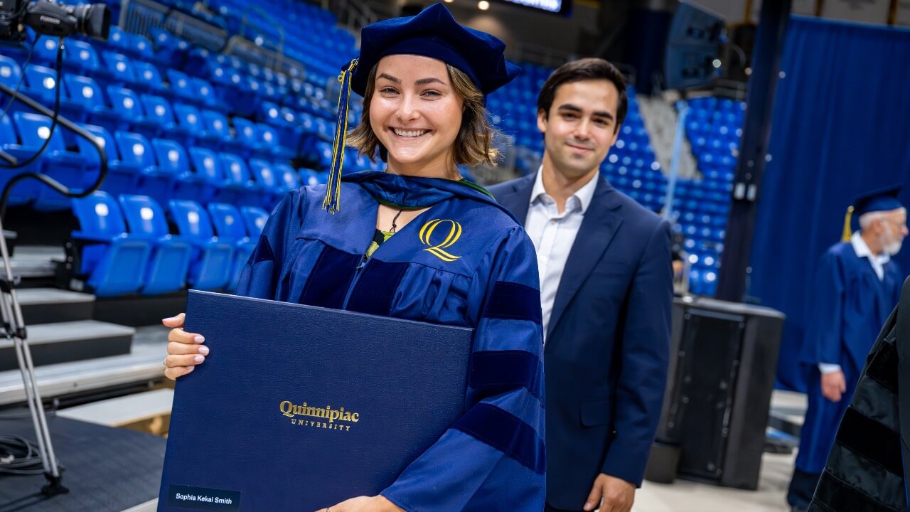 Graduate smiles and holds degree with loved one behind her after walking the Commencement stage
