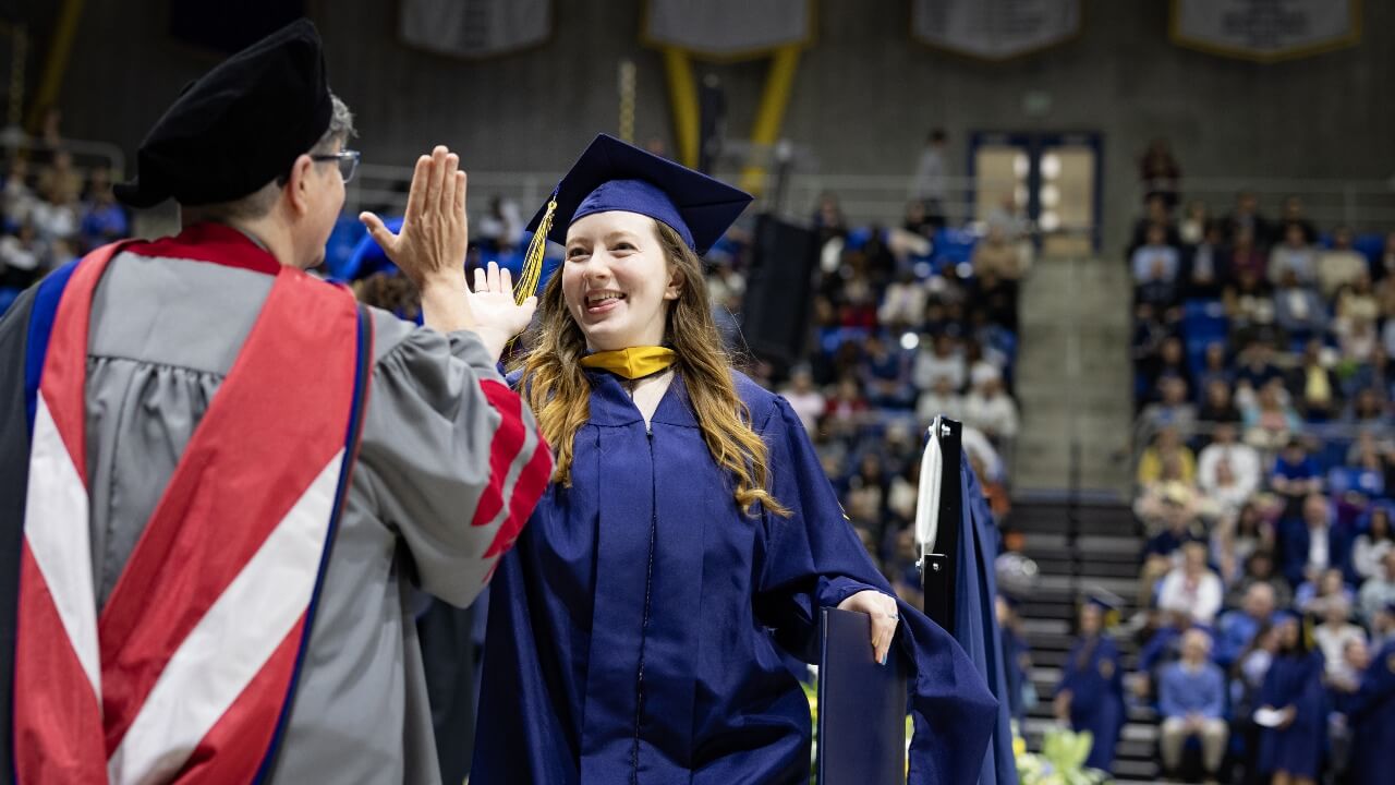 Graduate in cap and gown high-fives faulty member as they walk off stage.