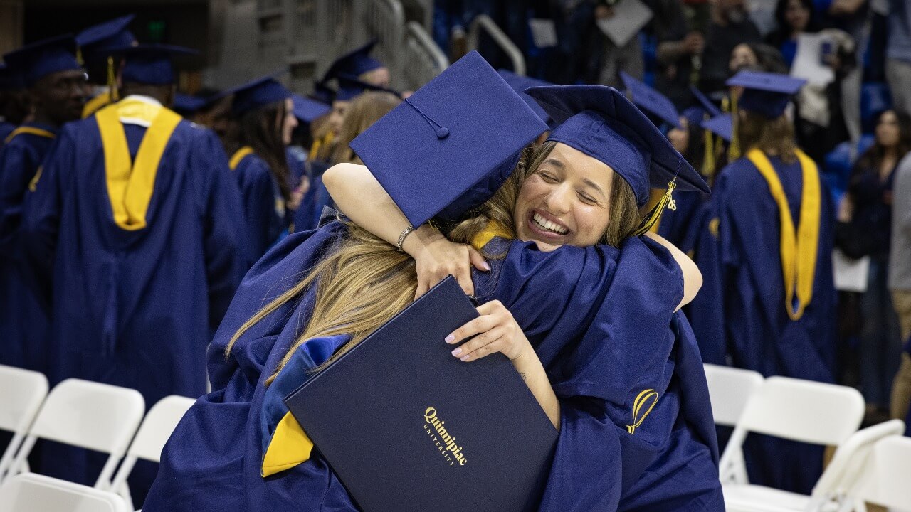 Two graduates hug while holding diplomas during ceremony
