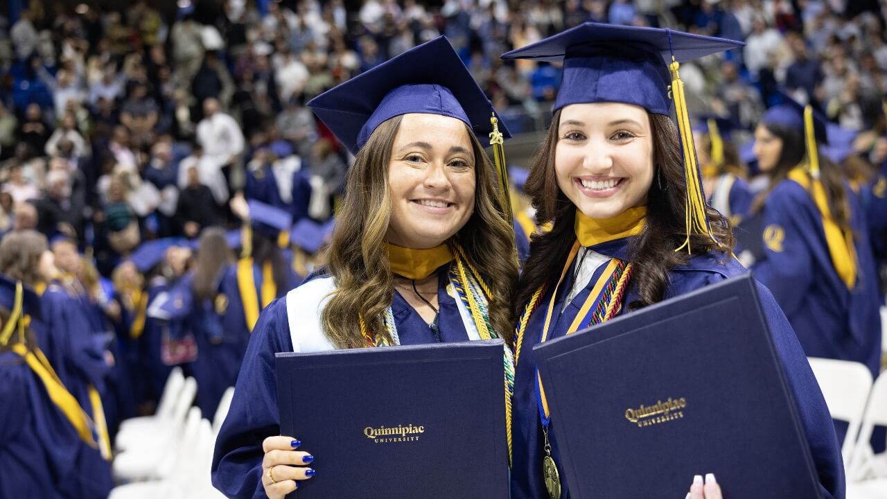 Two graduates smile and hold up their diplomas