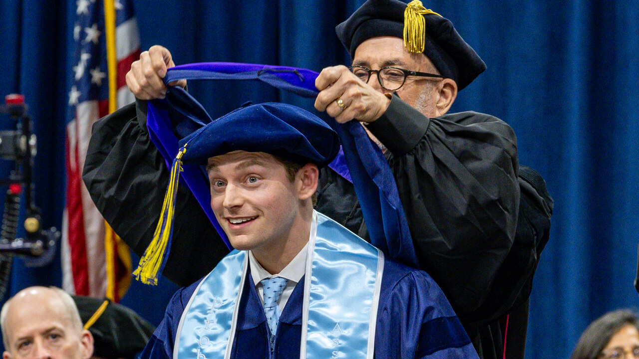 A graduate bends down as a professor drapes a doctoral hood on his head