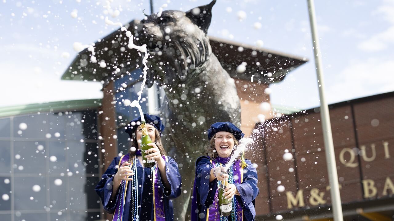 Two graduates pop champagne in front of the Bobcat statue
