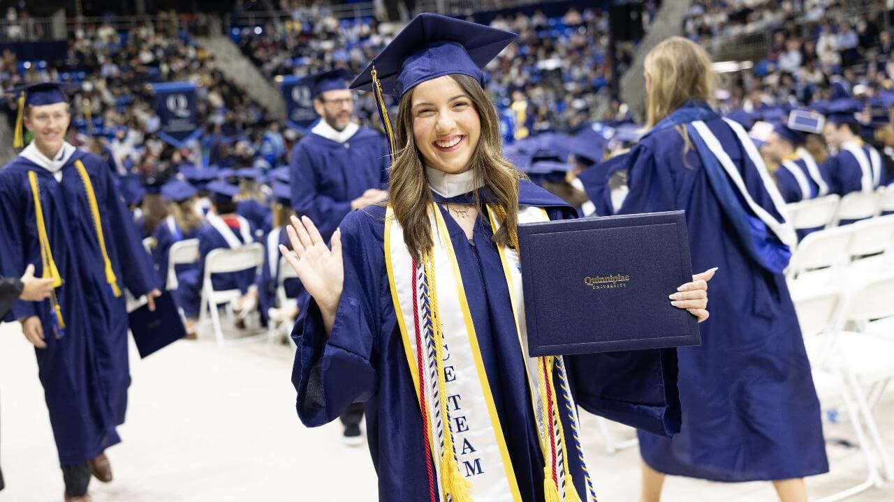 Graduate in cap and gown waves with their diploma.