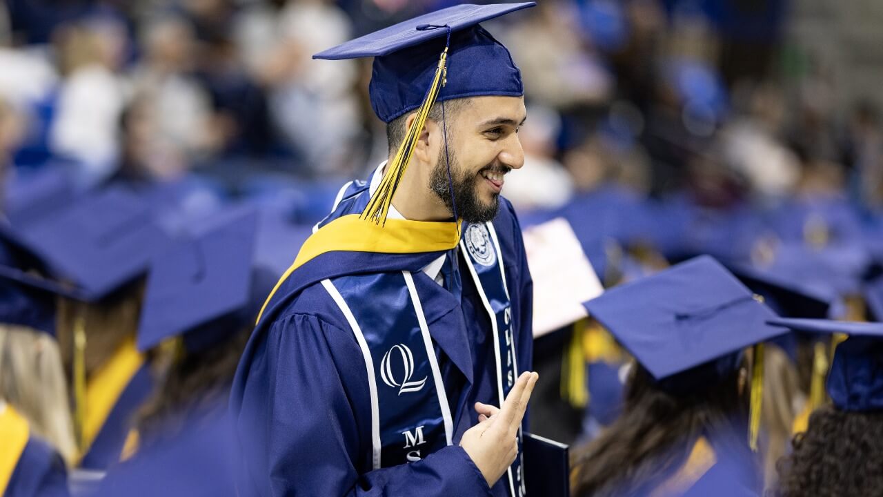 Graduate in cap and gown puts up a peace sign as they walk down the isle.