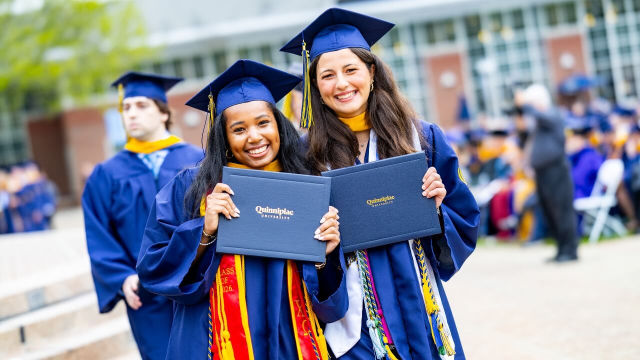 Two graduates in caps and gowns smile holding their diplomas.