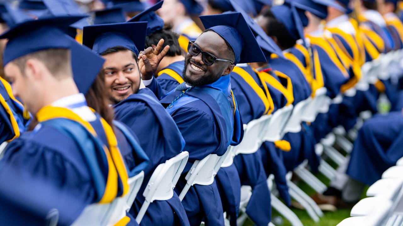 Graduate in cap and gown leans back in crowd to smile for a photo.