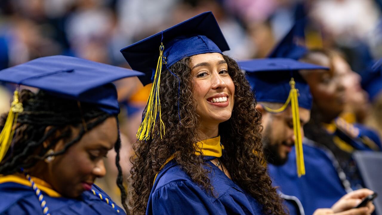 Graduate sits next to peers, all smiling as they enjoy the graduation ceremony