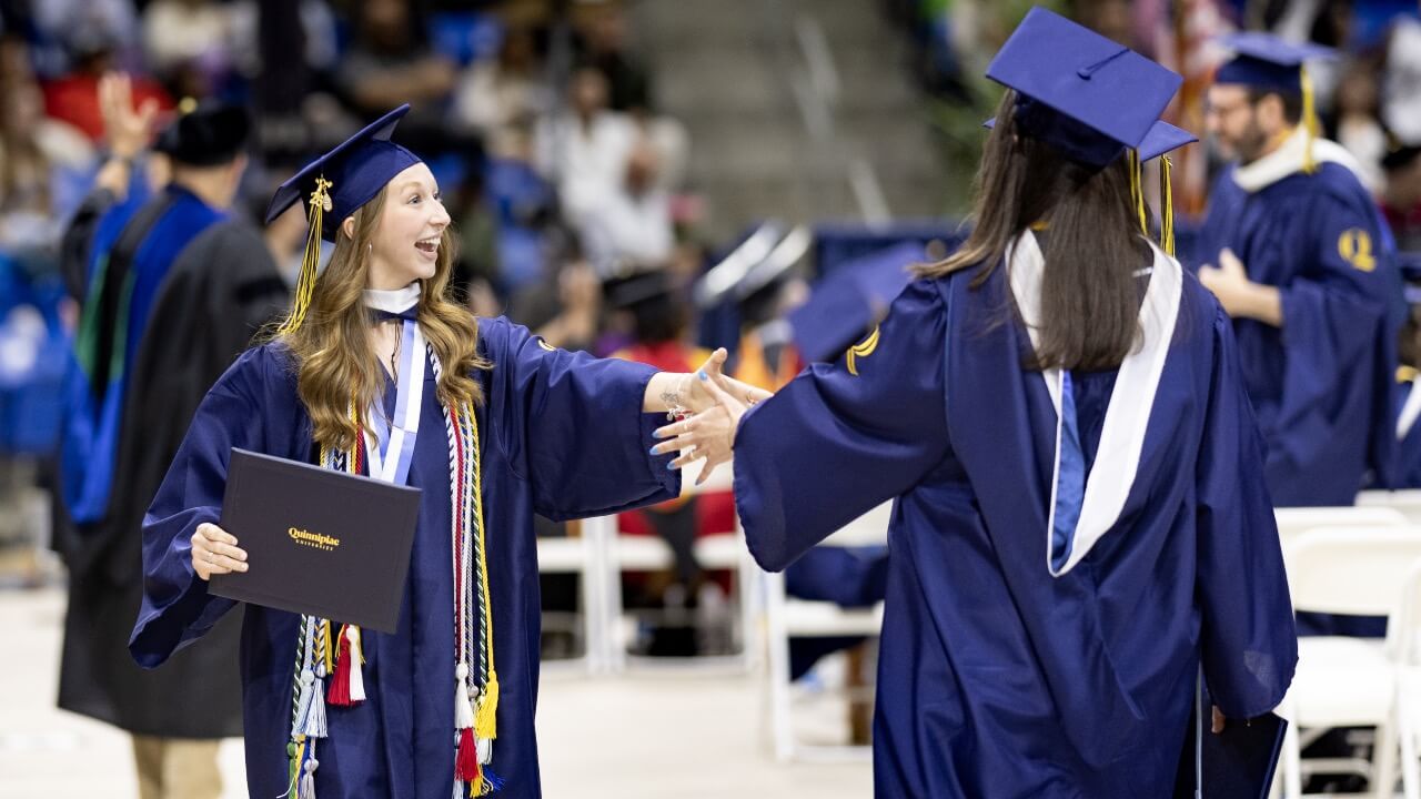 Two graduates smile and hold out hands as they pass eachother