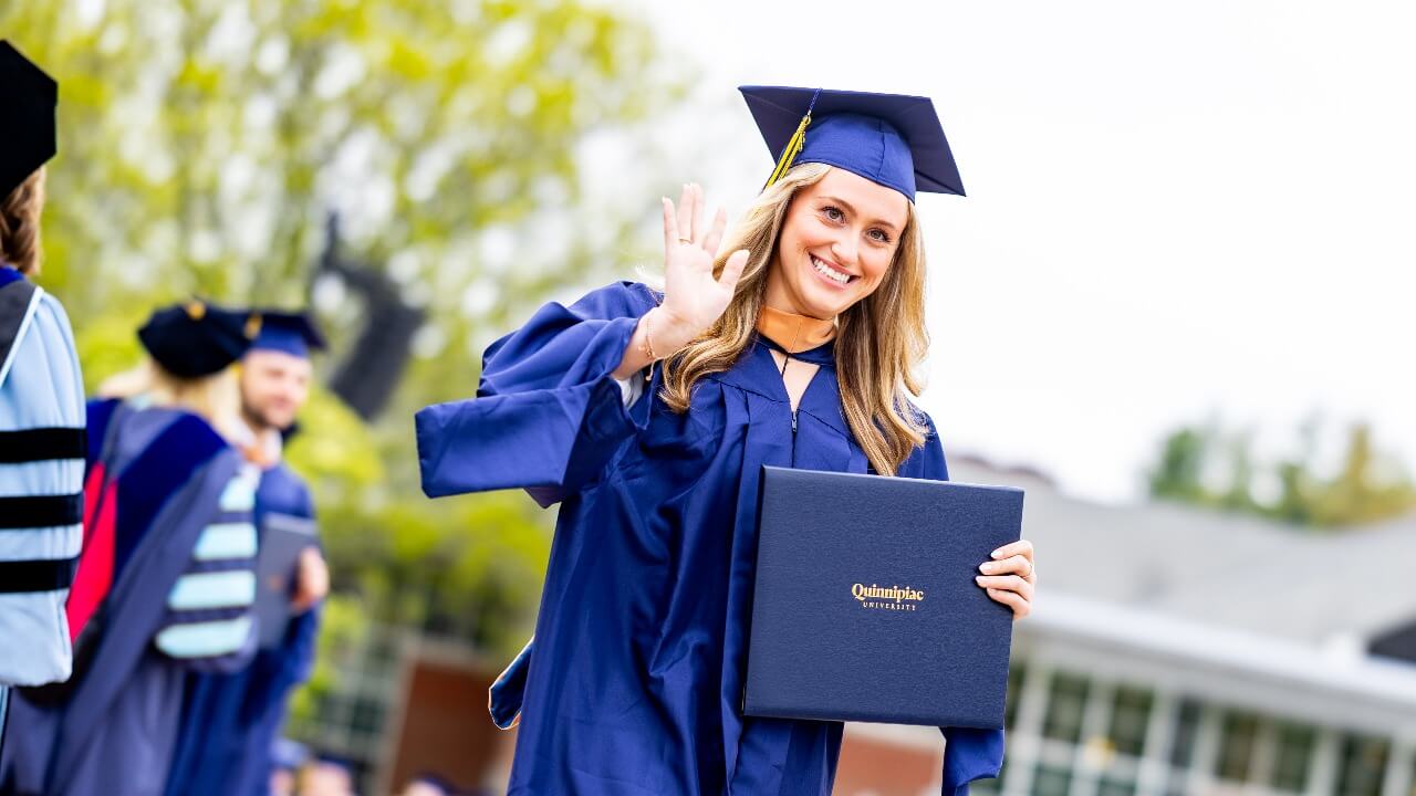 Graduate holding up her diploma with one hand and waving with the other hand