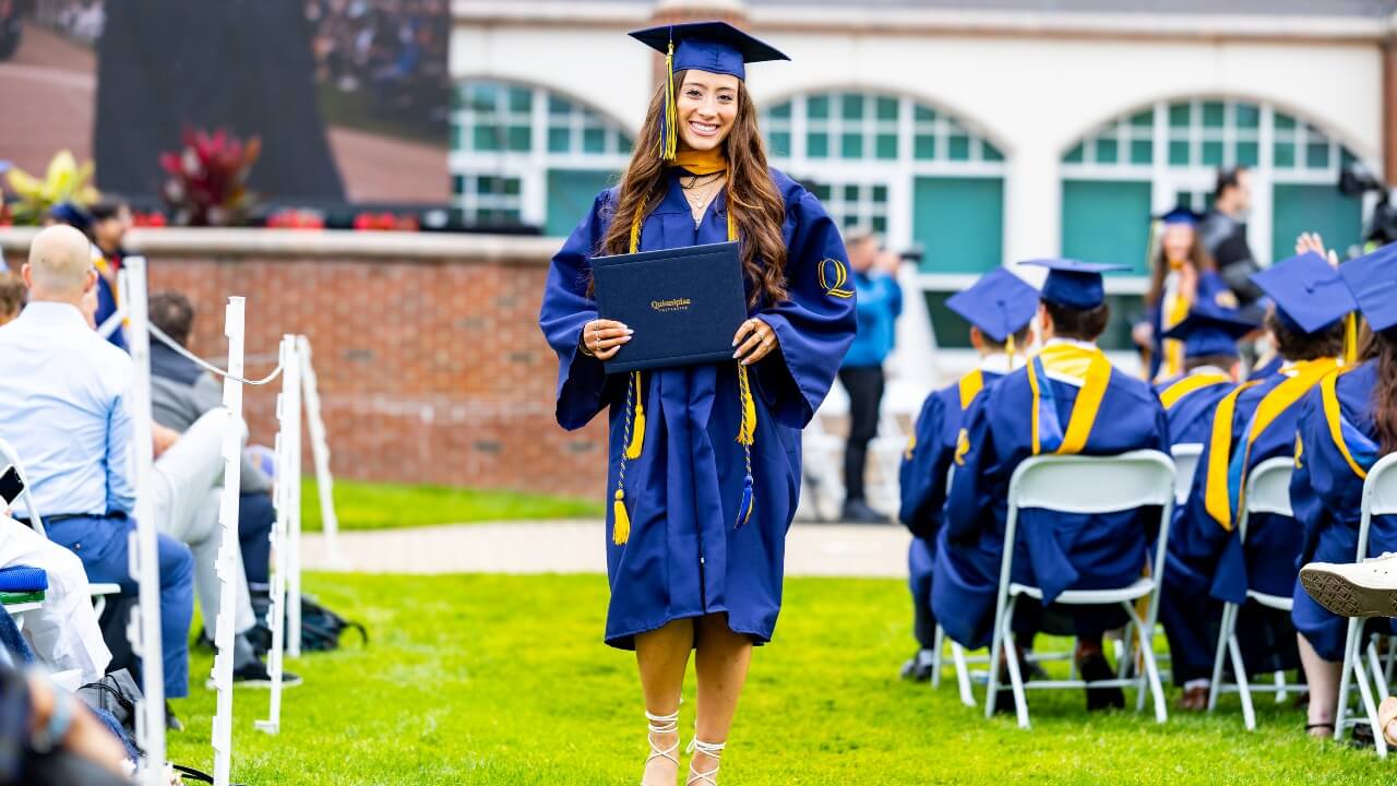 Graduate in cap and gown smiles holding their diploma as they walk down the isle.