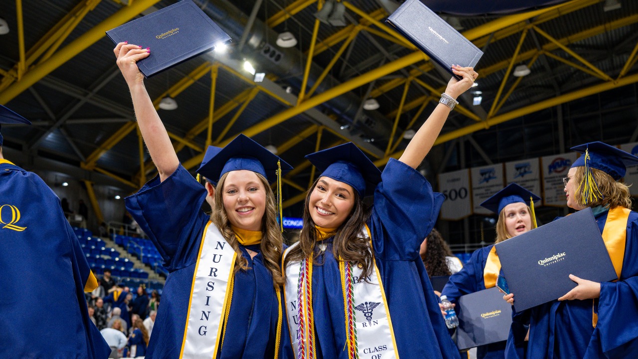 Two graduates dressed in cap and gown hold up their diplomas