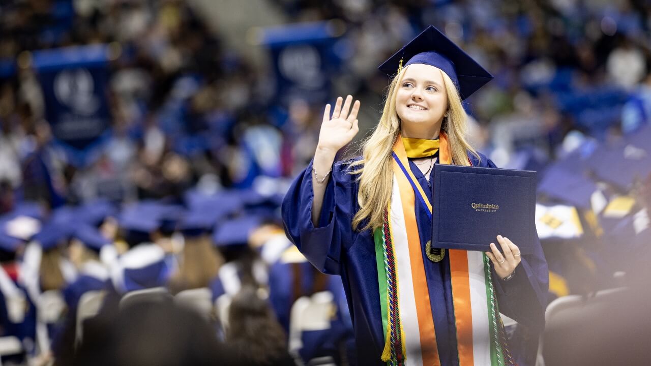 Graduate waves and looks at crowd while walking the stage at Commencement