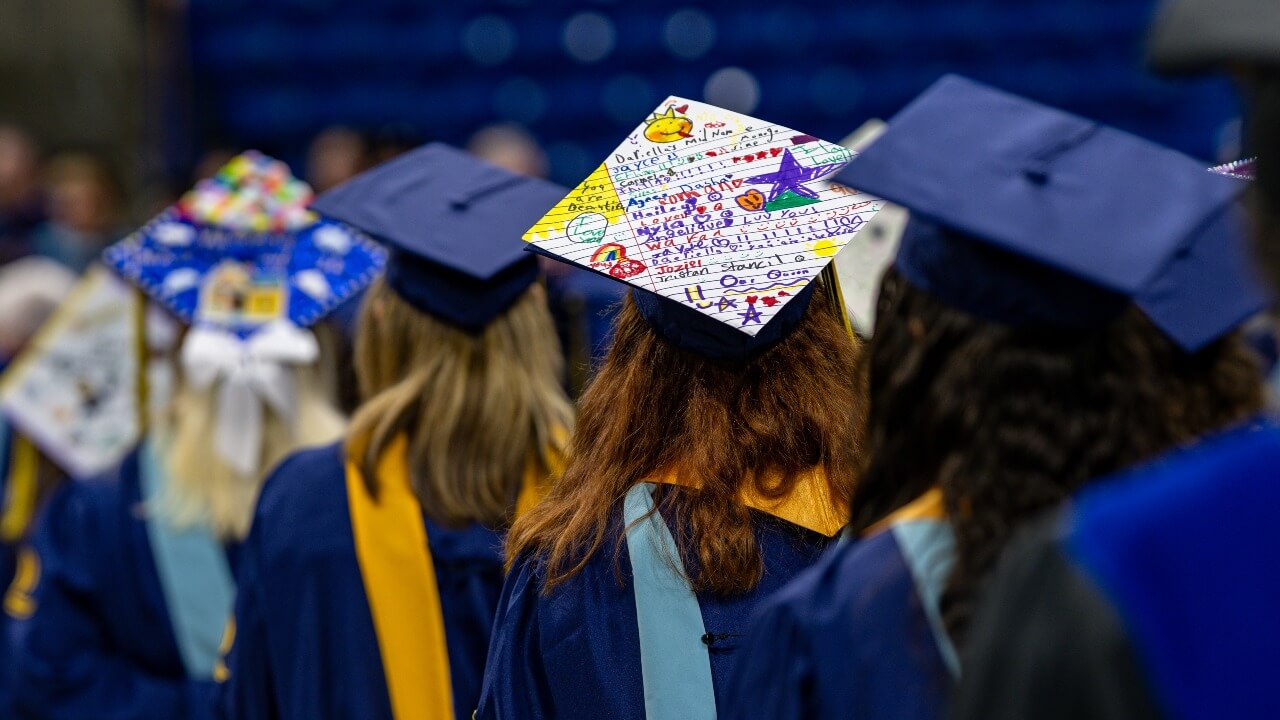 Graduates lined up facing away from the camera, showing their graduation cap designs