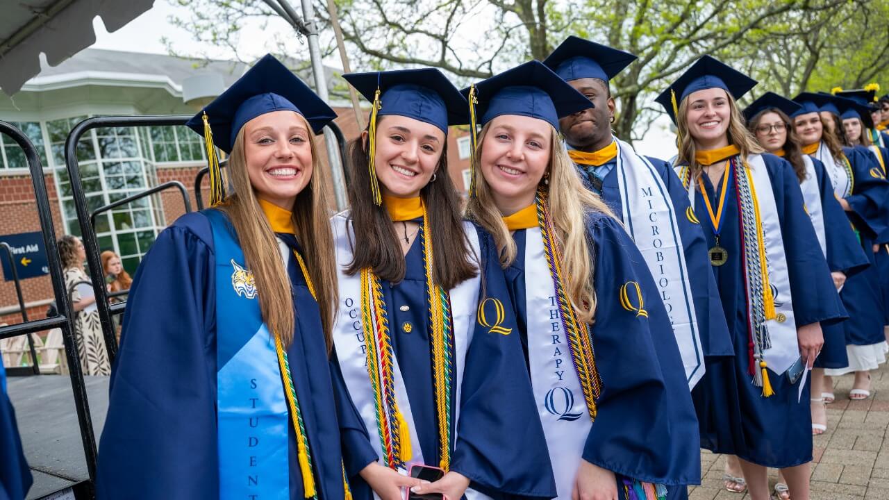 three graduating friends huddling together for a photo in front of a line of graduates