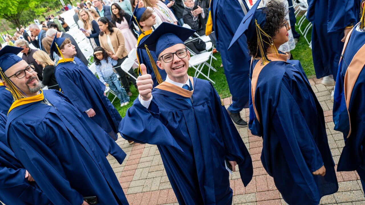 graduate in a crowd of people, giving a smile and a thumbs up to the camera