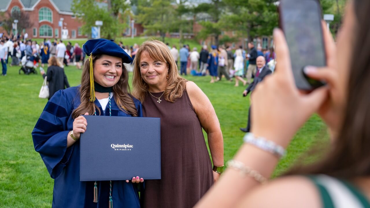 Individual taking a photo of a graduate with their diploma standing next to a loved one