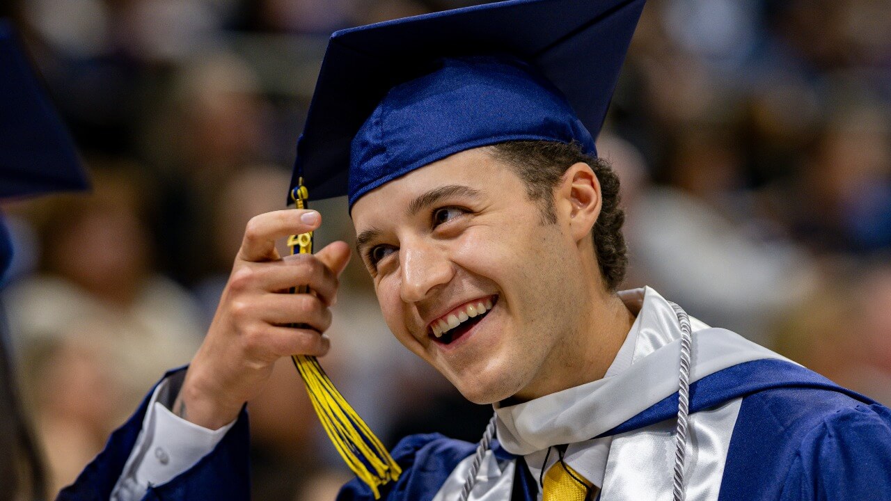 Graduate in cap and gown smiles and holds their tassel.