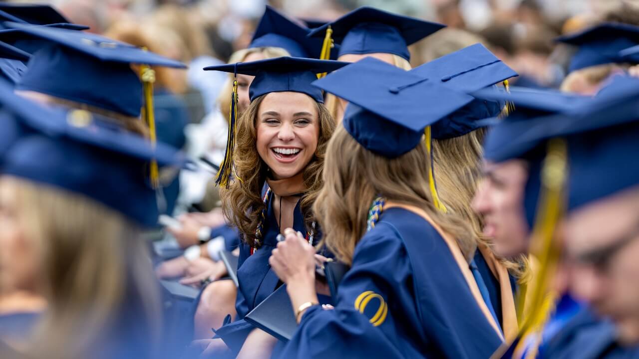 two graduates sitting in the crowd, laughing and clapping with each other