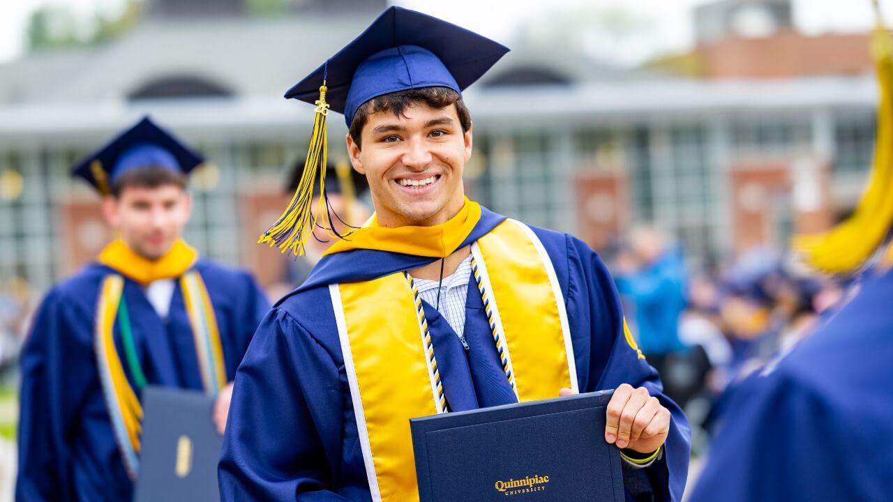 Graduate wearing regalia and a yellow stole while holding up diploma