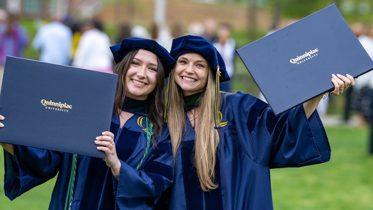 Two graduates in caps and gowns smile for a picture holding up their degrees.