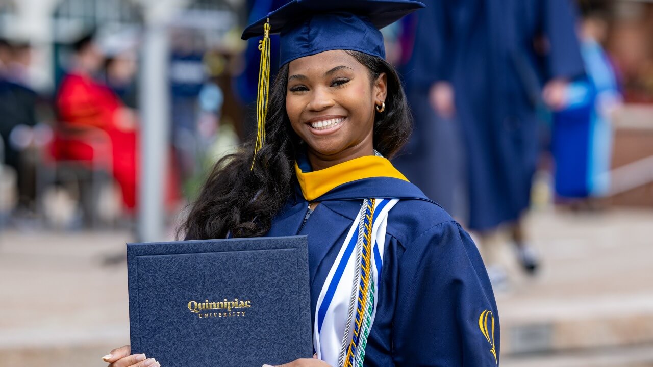 Graduate in cap and gown smiles for a photo holding their diploma.