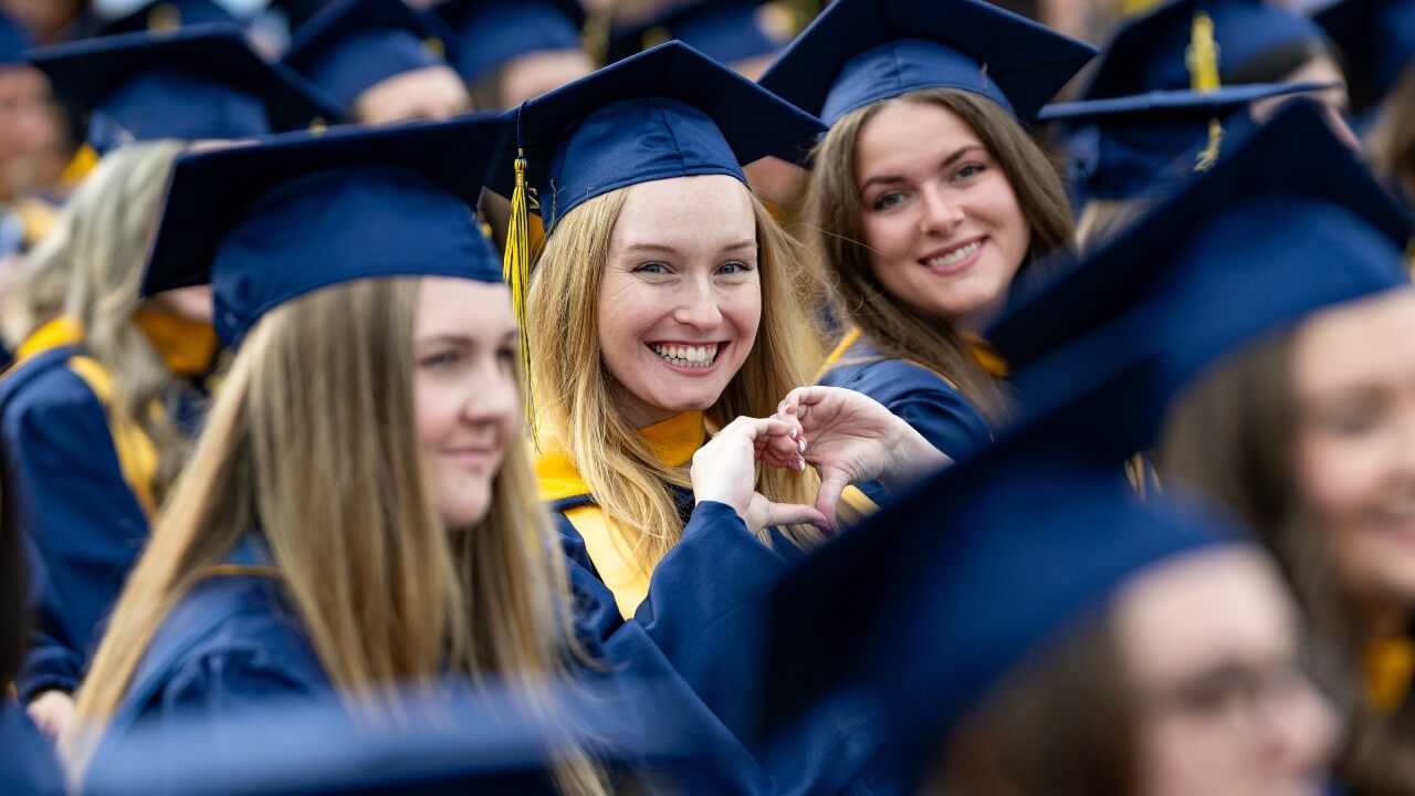 Graduates sitting in the crowd, one graduate is holding up a heart with her hands
