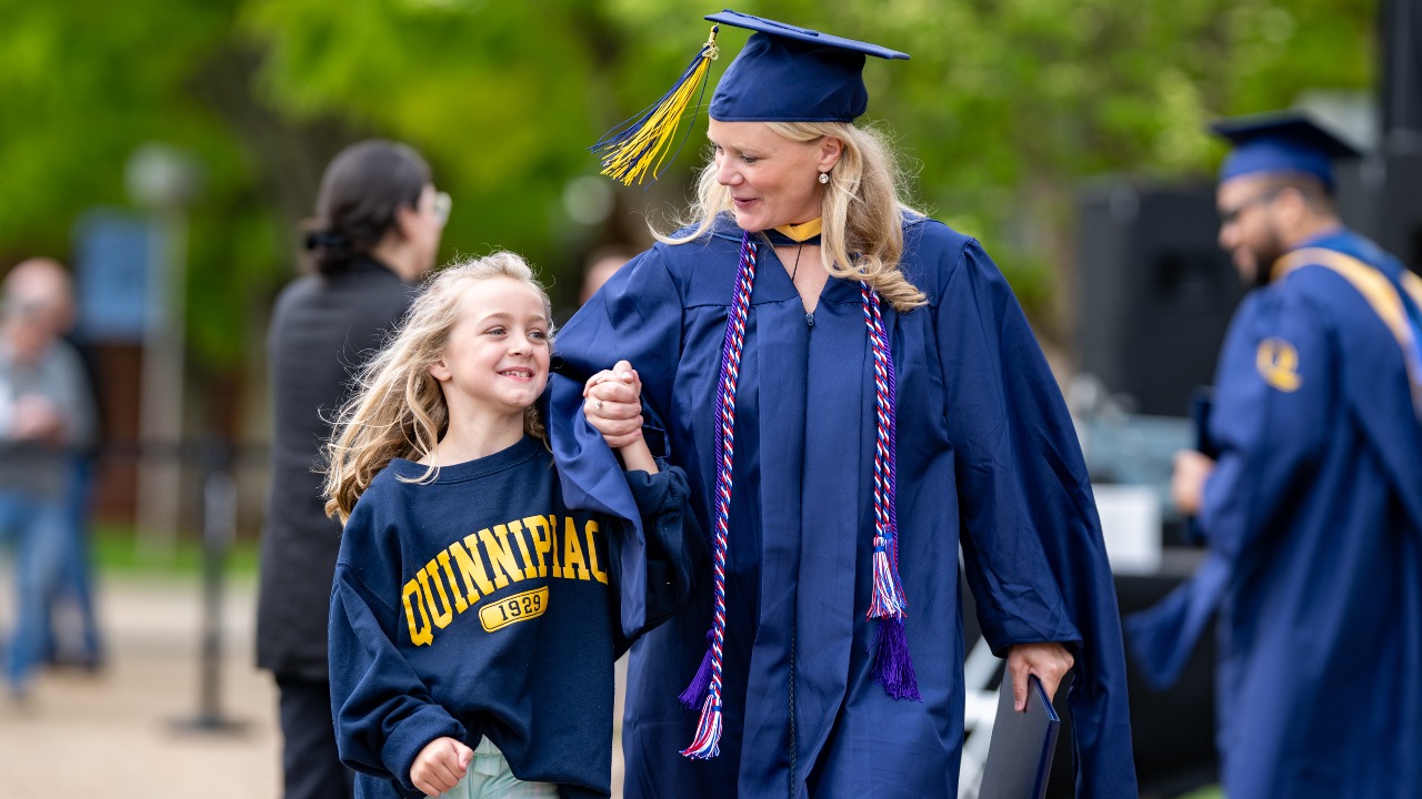a graduate in cap and gown holding hands and young child in an Quinnipiac sweatshirt