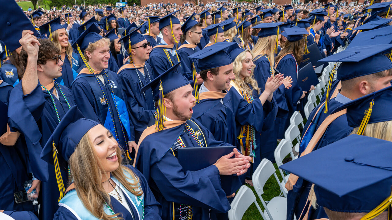 Hundreds of graduates in Commencement robes cheer and clap from their seats