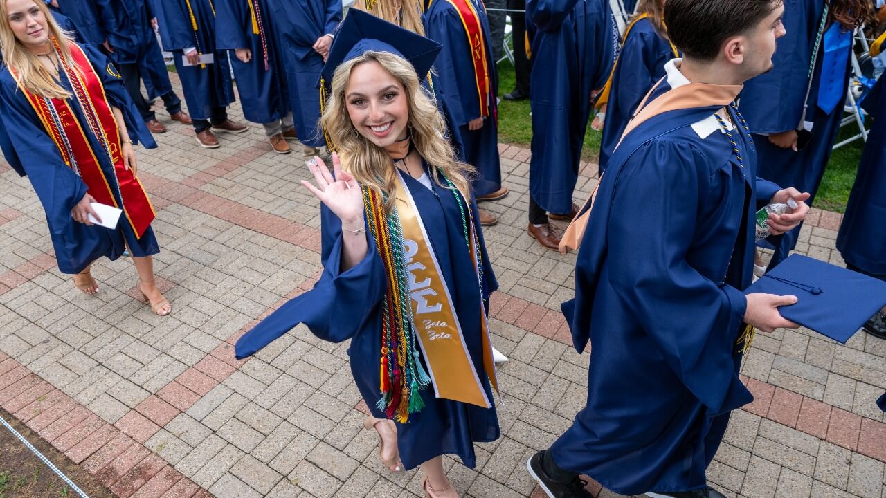 During walkout, graduate smiles and waves wearing cap and gown