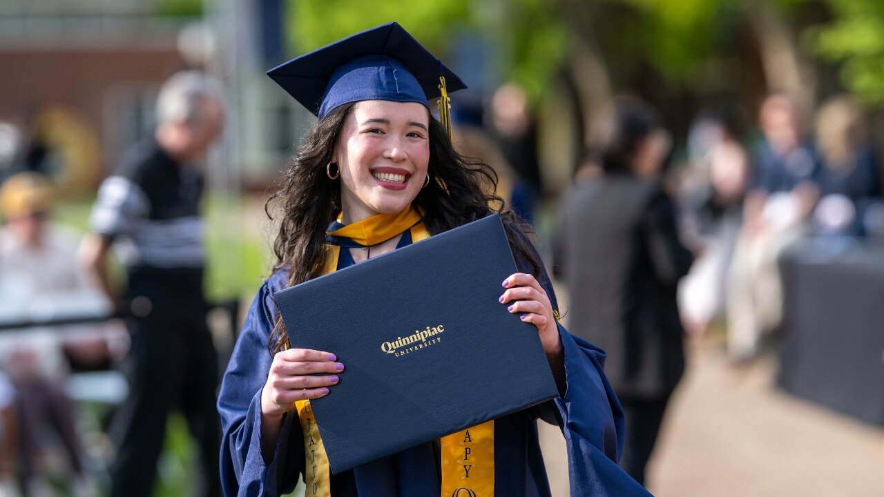 a joyful graduate smiling and proudly holding up their diploma for the camera