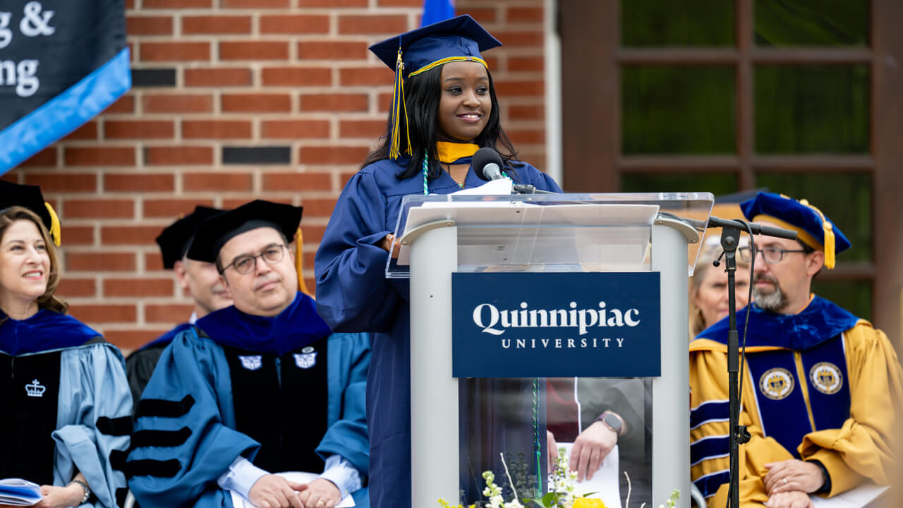 Margaret Gachau speaks at a podium with several administrators seated behind her listening