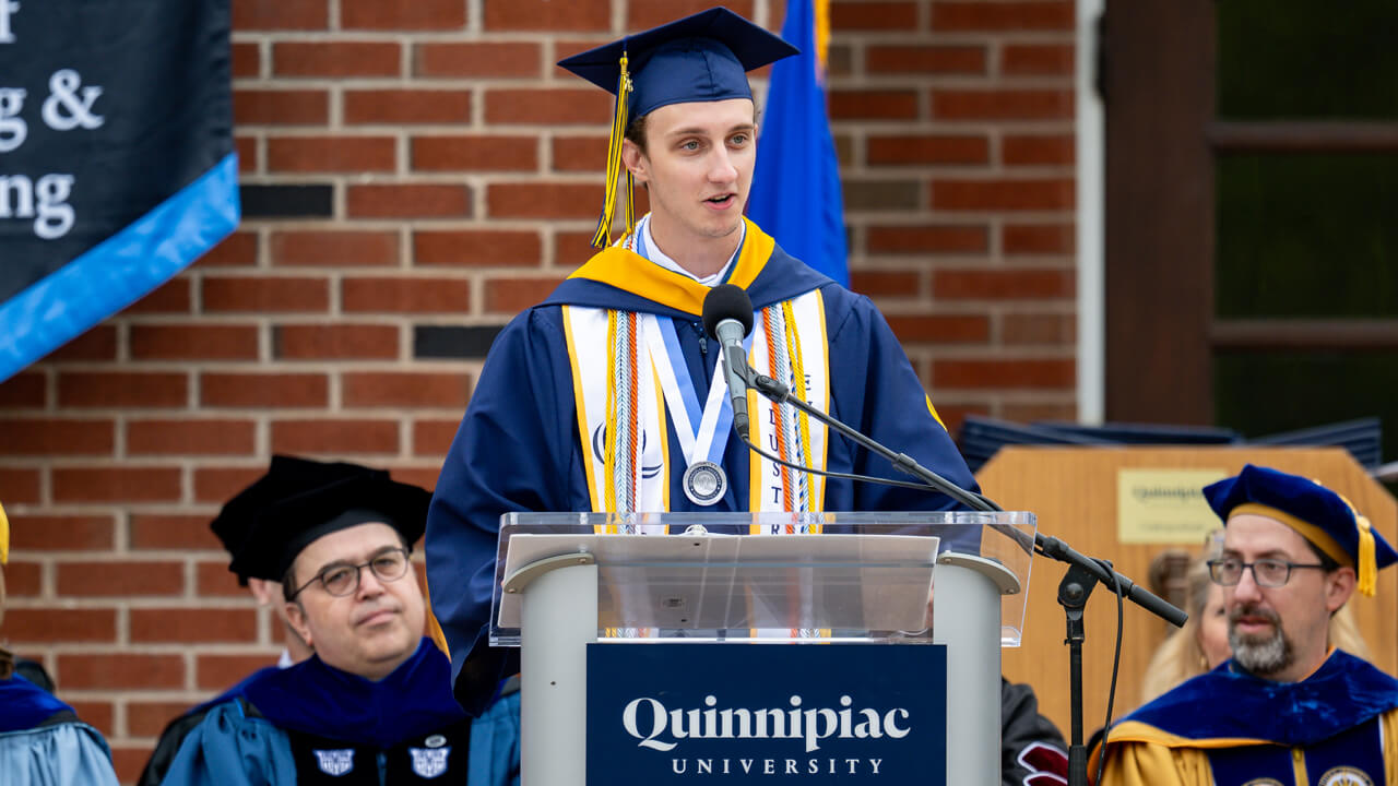 Gavin Grim wears Commencement robes, cap and several stoles and cords and speaks at a podium