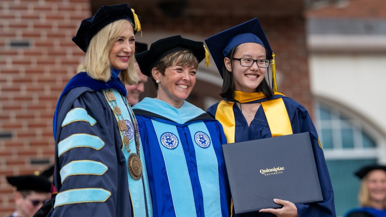 Graduate in cap and gown pose with photo with Marie Hardin and Monique Drucker