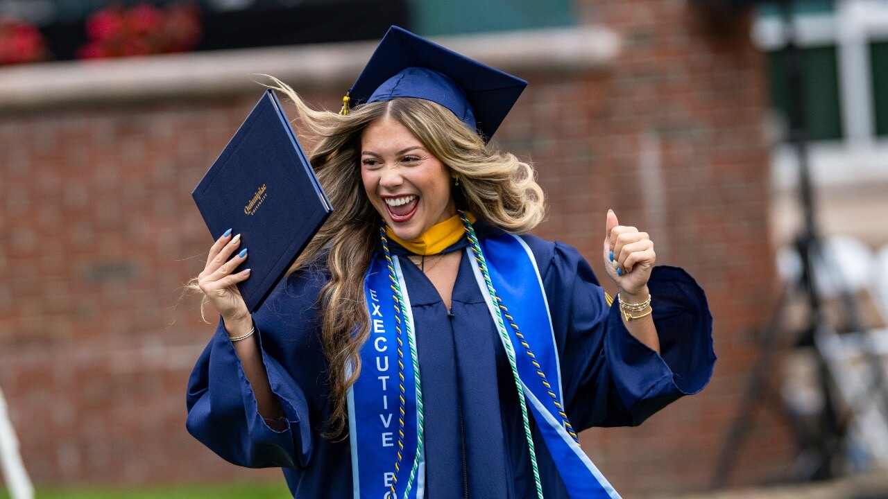 A graduate smiles broadly and cheers with arms up with a Quinnipiac diploma