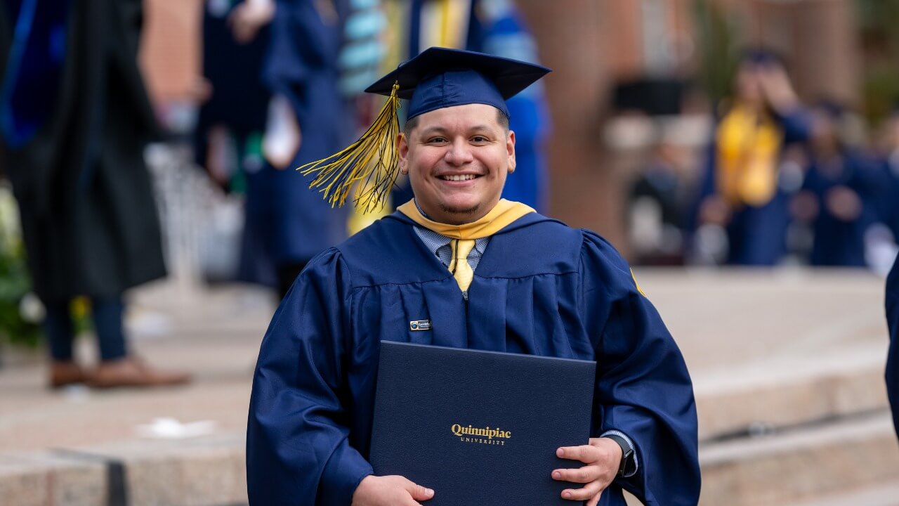 Graduate standing at the base of the library steps holding up diploma and smiling