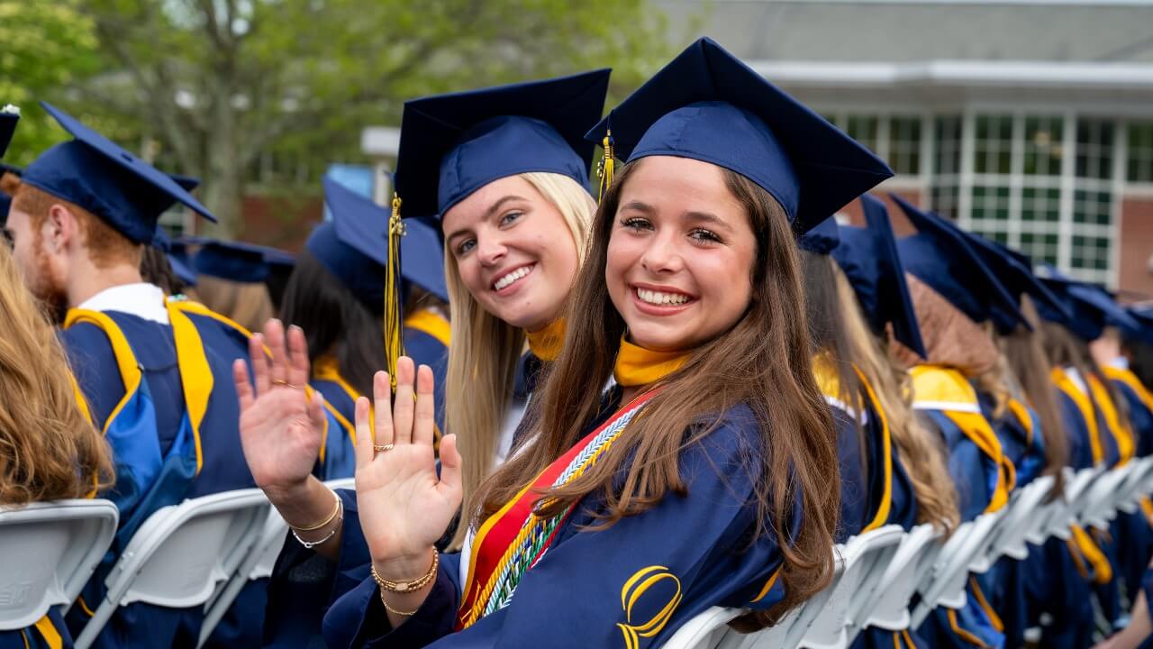 hundreds of graduates sitting in their seats while two look at the camera with a smile and wave