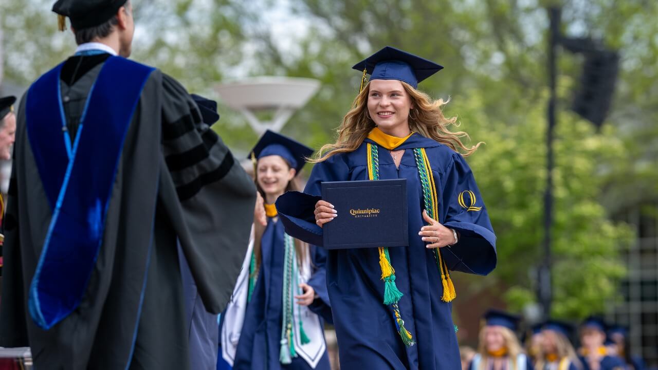 a graduate smiling and holding their diploma while walking in line