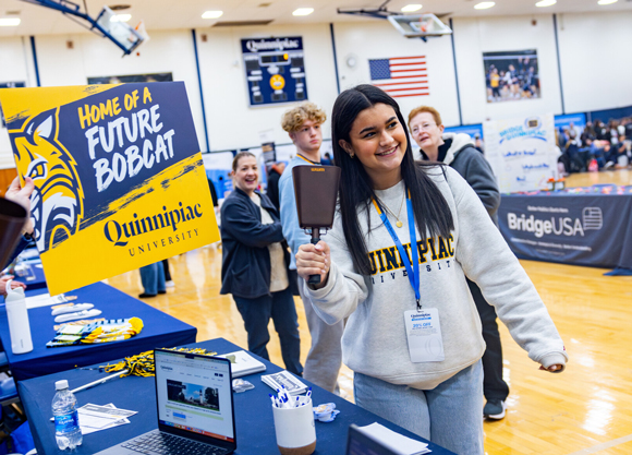 A new student in a Quinnipiac sweatshirt smiles and rings a cowbell in the rec center