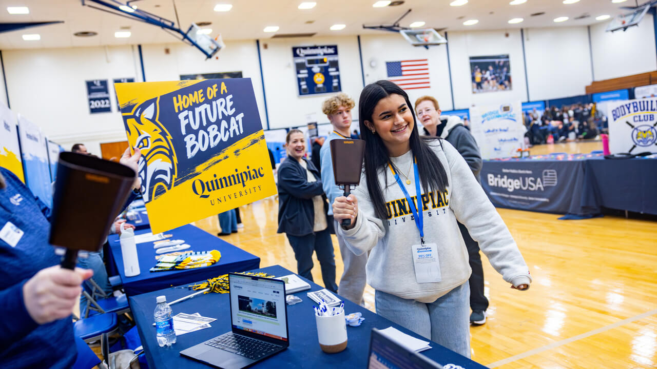 A new student in a Quinnipiac sweatshirt smiles and rings a cowbell in the rec center