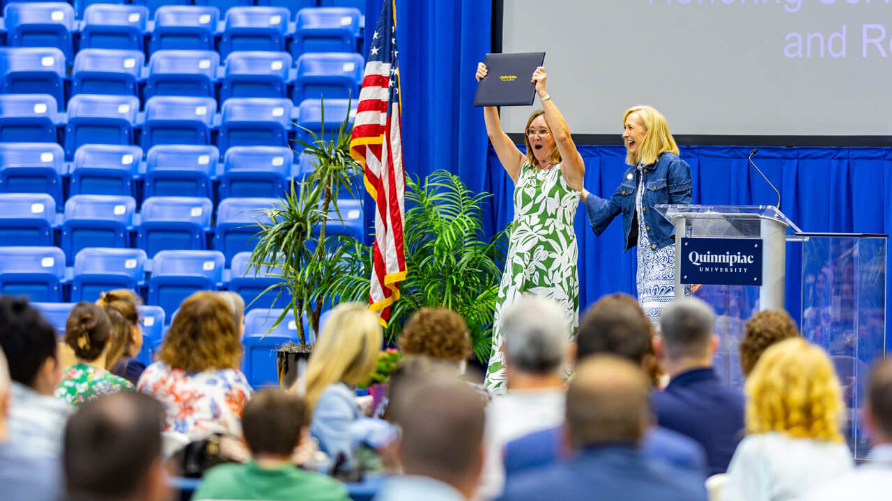 A Quinnipiac employee celebrates while holding a recognition certificate beside President Marie Hardin
