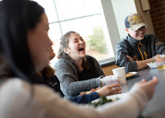 Three students laugh as they eat together in the Mount Carmel Campus dining hall