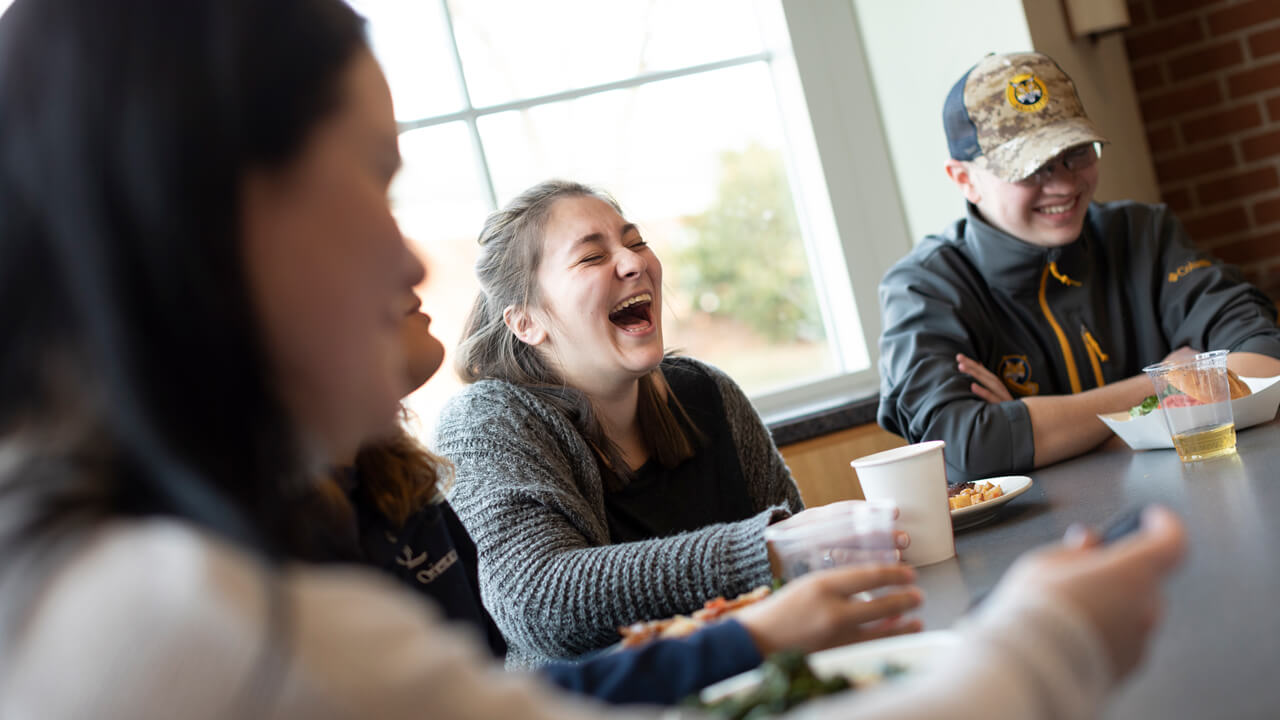 Three students laugh as they eat together in the Mount Carmel Campus dining hall